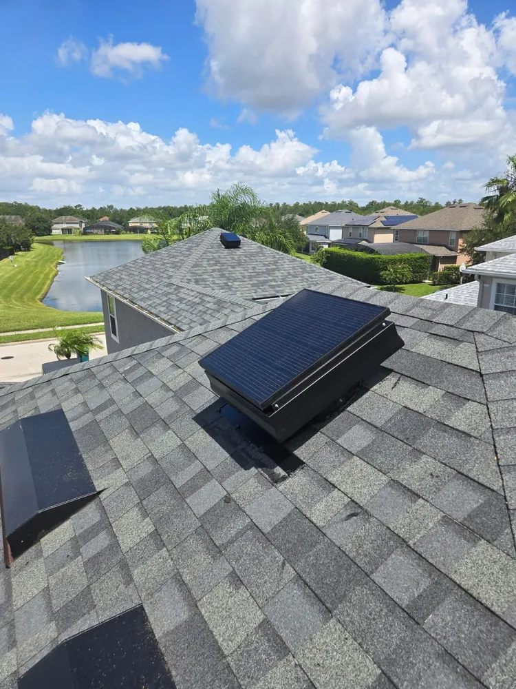Solar attic fan on a grey shingle roof overlooking a neighborhood lake